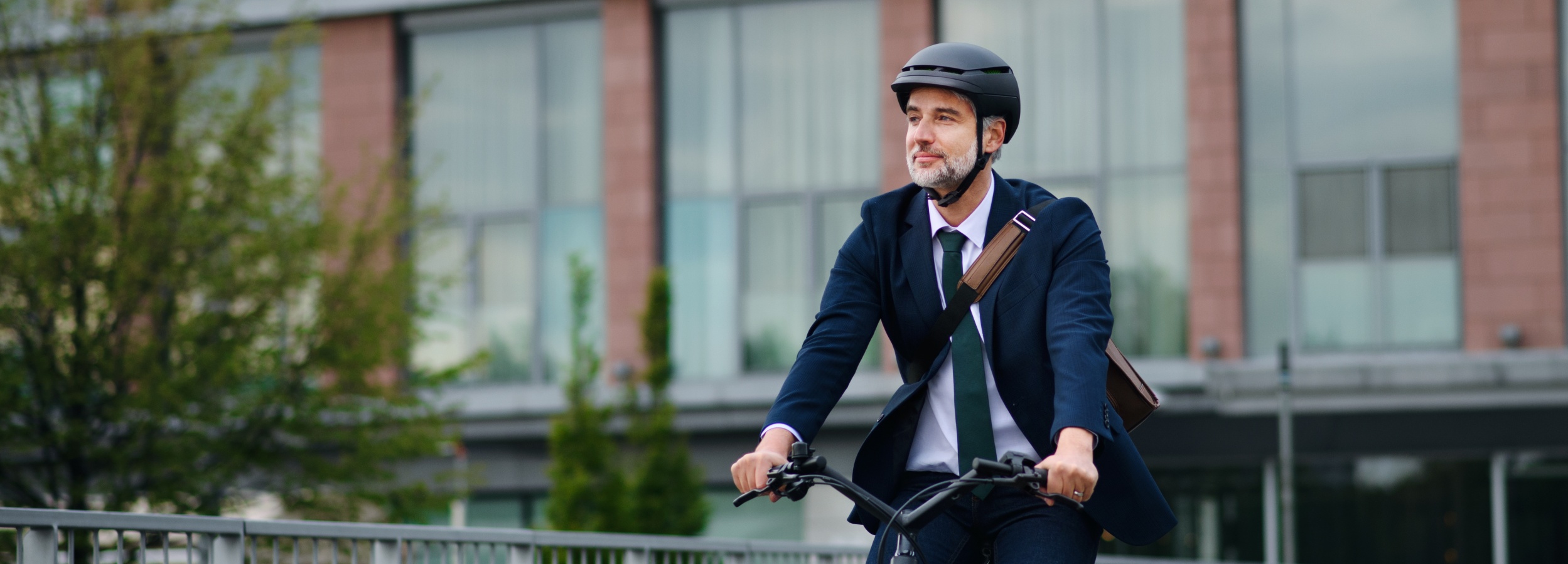 Ein grauhaariger Mann in Anzug und Helm fährt mit einem Fahrrad durch die Stadt.

Das Bild zeigt einen erwachsenen Mann mittleren Alters, der einen dunklen Anzug, ein weißes Hemd und eine grüne Krawatte trägt und auf einem Fahrrad sitzt. Er hat graue Haare und einen Bart und trägt einen schwarzen Helm. Seine Hände halten den Lenker des Fahrrads fest, und er blickt lächelnd nach rechts. Auf seiner rechten Schulter trägt er eine braune Umhängetasche. Das Fahrrad selbst ist dunkel und scheint ein moderneres Modell zu sein, mit einem Scheinwerfer vorne. Der Mann befindet sich auf einer Brücke oder einem erhöhten Weg, der von einem metallenen Geländer gesäumt wird. Im Hintergrund ist ein großes Gebäude mit roten Ziegelsteinfassaden und großflächigen Fenstern zu sehen. Vor dem Gebäude stehen Bäume mit hellem, grünem Laub. Die Atmosphäre des Bildes ist optimistisch und dynamisch, was durch den Blick des Mannes und seine Kleidung, die eine Mischung aus professioneller Eleganz und aktiver Mobilität darstellt, unterstrichen wird. Die Beleuchtung scheint diffus zu sein, was auf einen bewölkten Tag oder eine spätere Tagesstunde hindeutet, aber die Szene wirkt dennoch lebendig.