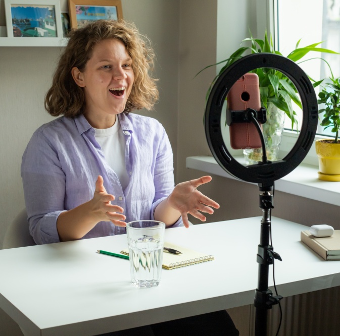 Das Bild zeigt eine junge Frau mit lockigen, hellbraunen Haaren, die in die Kamera blickt, während sie spricht. Sie trägt ein hellviolettes Hemd über einem weißen T-Shirt. Ihre Augenbrauen sind leicht hochgezogen, und ihr Mund ist weit geöffnet, was auf Begeisterung oder eine lebhafte Erzählung hindeutet. Ihre Hände sind erhoben und ausgestreckt, als ob sie ihre Worte mit Gesten unterstreicht. Sie sitzt an einem weißen Schreibtisch. Vor ihr steht ein Glas mit einer klaren Flüssigkeit, wahrscheinlich Wasser, das durch die Reflexionen und Blasen belebt wirkt. Daneben liegen ein Stift und ein Notizbuch mit Spiralbindung. Im Hintergrund ist ein Ringlicht mit einem Smartphone in der Mitte aufgestellt, das eindeutig für eine Aufnahme oder einen Livestream gedacht ist. Hinter dem Tisch, in der Nähe eines Fensters, stehen Pflanzen in hellgelben und anderen grünen Töpfen, die etwas Leben in die Szene bringen. Auf einem Regal über der Frau sind mehrere gerahmte Bilder zu sehen. Die allgemeine Atmosphäre ist hell und freundlich, mit einer leichten Neigung zur Konzentration und Begeisterung der Frau. Die Stimmung ist lebendig und kommunikativ, was darauf hindeutet, dass die Frau entweder Inhalte erstellt, unterrichtet oder sich mit anderen austauscht.