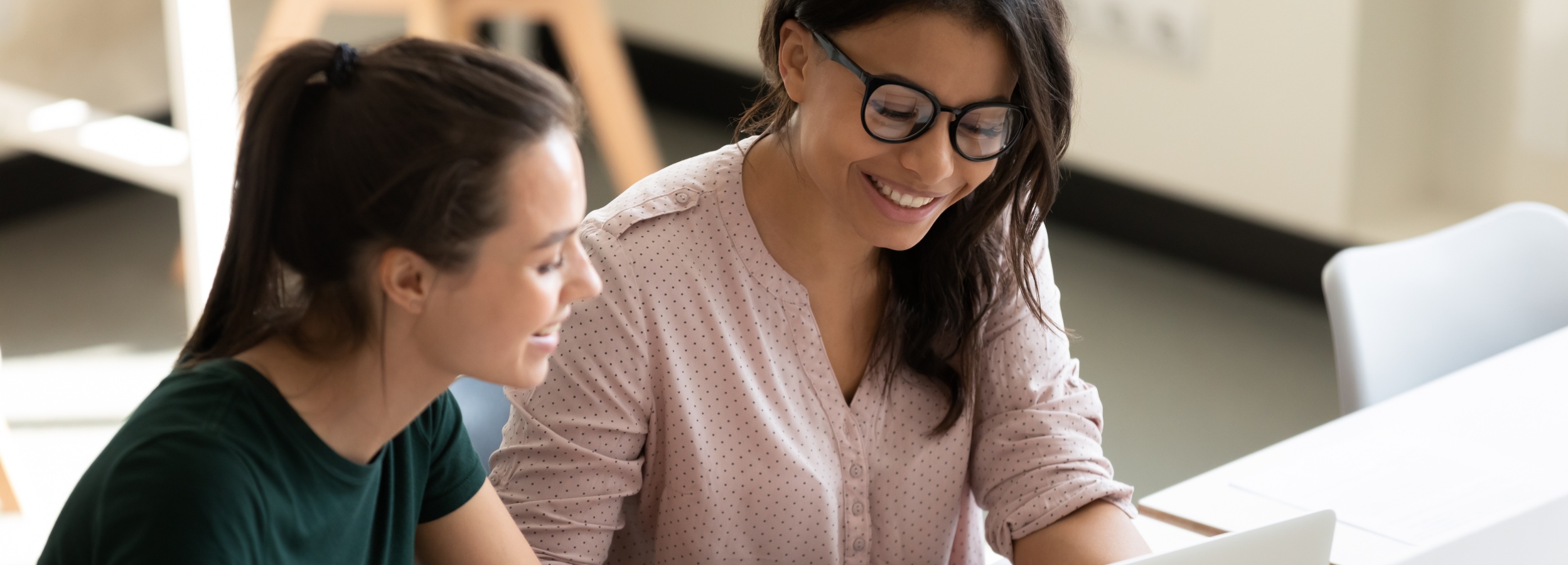 Das Bild zeigt zwei lächelnde Frauen, die gemeinsam an einem Laptop arbeiten und eine positive und kollaborative Arbeitsatmosphäre ausstrahlen.
Im Detail sieht man zwei Frauen, die eng nebeneinander an einem weißen Tisch sitzen. Die Frau links trägt ein dunkelgrünes T-Shirt und hat ihre braunen Haare zu einem Pferdeschwanz gebunden. Sie lacht leicht und schaut auf den Laptop, den die andere Frau hält. Die Frau rechts trägt eine hellrosa Bluse mit kleinen weißen Punkten und eine Brille. Sie lächelt breit und scheint konzentriert auf den Laptop zu sein. Ihr dunkles Haar fällt ihr über die Schultern. Der Laptop ist im Bildausschnitt nur teilweise sichtbar. Im Hintergrund sieht man unscharf die Beine von Stühlen und einen hellen Raum. Die allgemeine Atmosphäre ist freundlich, positiv und konzentriert. Die Stimmung wird durch die lächelnden Gesichter der Frauen und die Nähe zueinander vermittelt. Das Bild strahlt Zusammenarbeit und positive Arbeitsbeziehungen aus.