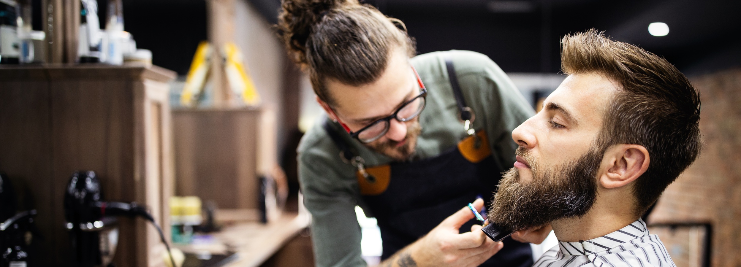 Ein Friseur trimmt den Bart eines Kunden in einem Barbershop.

Das Bild zeigt einen Mann mit einem Dutt und Brille, der als Friseur in einem Barbershop tätig ist. Er trägt eine grüne Arbeitskleidung und eine schwarze Schürze. Er steht über einem Kunden und trimmt dessen Bart mit einer Schere und einem Kamm. Der Kunde hat einen vollen, gepflegten Bart und trägt einen Umhang mit schwarzen und weißen Streifen. Die Atmosphäre wirkt professionell und entspannt. Im Hintergrund sind Regale mit Friseurprodukten und Geräten zu sehen, die den Eindruck eines gut ausgestatteten Barbershops verstärken. Die Stimmung ist ruhig und konzentriert, da der Friseur sorgfältig an der Bartpflege arbeitet.