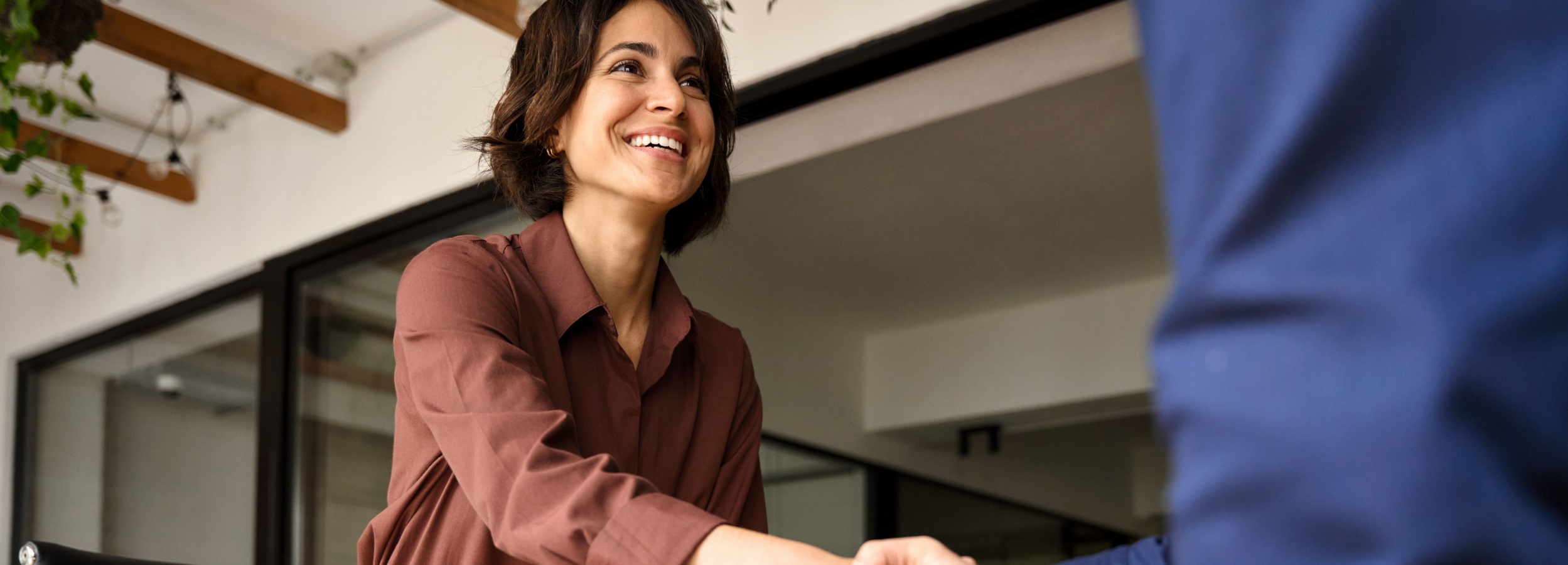 Ein freundliches Lächeln und ein Händedruck signalisieren eine positive geschäftliche Übereinkunft.

Das Bild zeigt eine junge Frau mit dunklem, schulterlangem Haar, die ein burgunderrotes Hemd trägt und herzlich lächelt, während sie scheinbar jemandem die Hand schüttelt. Nur der Arm und ein Teil des Oberkörpers der anderen Person, bekleidet mit einem blauen Jackett, sind im Bild sichtbar. Der Hintergrund ist etwas verschwommen, aber man erkennt Holzbalken an der Decke und eine Fensterfront mit Blick nach draußen, wo grüne Pflanzen hängen. Die Atmosphäre wirkt warm und professionell, und die Stimmung ist positiv und einladend.