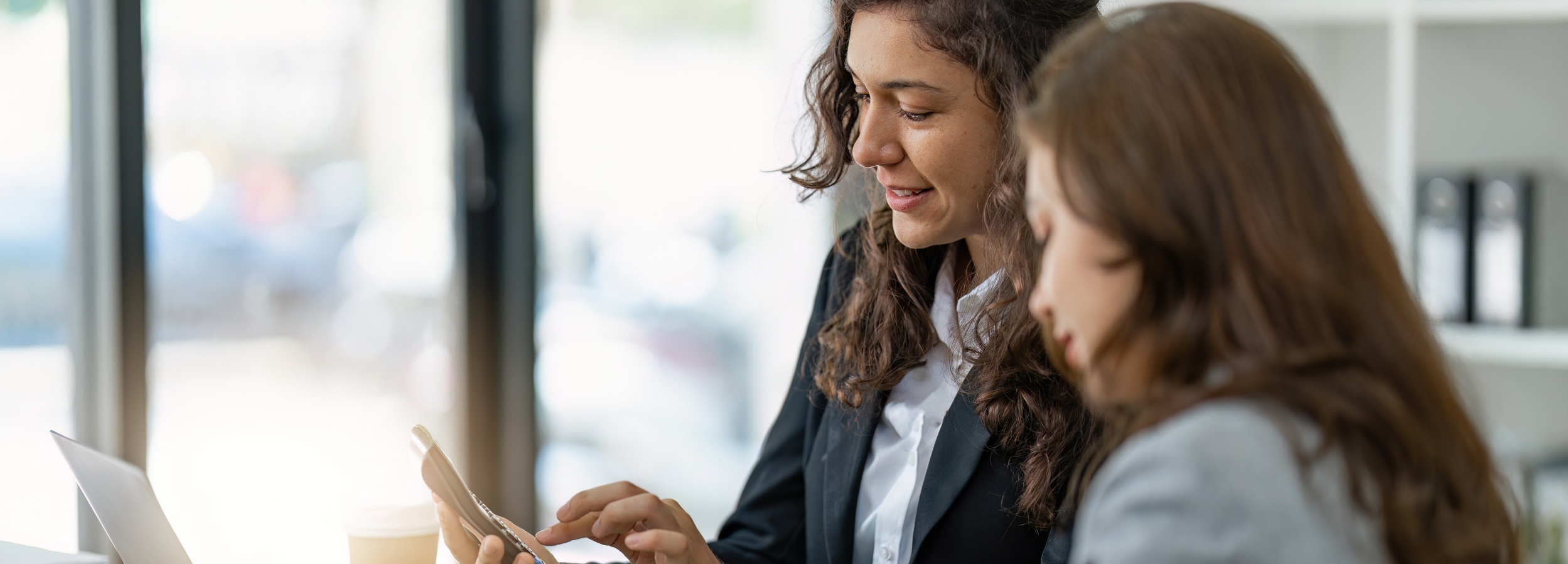 Zwei junge Geschäftsfrauen in formeller Kleidung besprechen ein digitales Tablet in einem hellen, modernen Büro.

Das Bild zeigt zwei Frauen, die wahrscheinlich in einem geschäftlichen Kontext stehen. Die Frau im Vordergrund trägt einen schwarzen Blazer über einem weißen Hemd, ihr dunkles, lockiges Haar fällt ihr über die Schultern. Sie blickt auf ein digitales Tablet, das sie in den Händen hält und bedient. Ein Lächeln umspielt ihren Mund, was auf eine positive oder interessierte Haltung hindeutet. Neben ihr steht eine zweite Frau mit glattem, braunem Haar und einem hellgrauen Blazer, die ebenfalls auf das Tablet schaut.

Im Hintergrund ist ein Büroambiente zu erkennen. Helle Fenster lassen viel Tageslicht herein, und im Hintergrund deutet ein Regal auf eine Büroumgebung hin. Ein Laptop und ein Coffee-to-go-Becher sind ebenfalls auf dem Tisch zu sehen. Die Atmosphäre wirkt professionell und konzentriert, aber dennoch entspannt genug für ein offenes Gespräch oder eine Zusammenarbeit. Das Bild vermittelt eine Stimmung von moderner Arbeitsumgebung, Teamarbeit und positiver Interaktion.